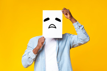 African american guy hiding face behind paper with sad emoticon