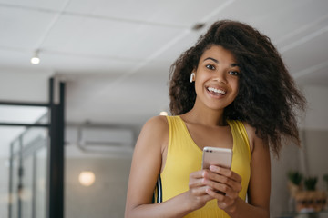 Beautiful excited African American woman holding cellphone, ordering food, shopping online on Black Friday. Emotional curly haired hipster girl listening music, laughing, using wireless earphones 