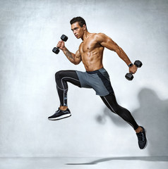 Sporty man jumping with dumbbells. Photo of active man with naked torso on grey background. Dynamic...