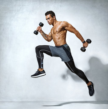 Sporty man jumping with dumbbells. Photo of active man with naked torso on grey background. Dynamic movement. Side view. Sport and healthy lifestyle