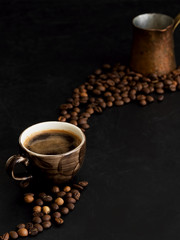 Cup with dark espresso on a black background. Roasted coffee beans are located around a cup of coffee. In the background is a copper cezve. Close-up.