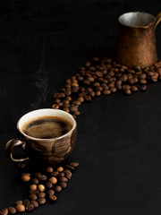 A cup with dark espresso on a black background, steam rises above the cup. Roasted coffee beans are located around a cup of coffee. In the background is a copper cezve. Close-up.