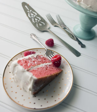 Piece Of Pink Layer Cake With White Frosting And Fresh Raspberry Garnish. Shown With Utensils And Fancy Plate. 