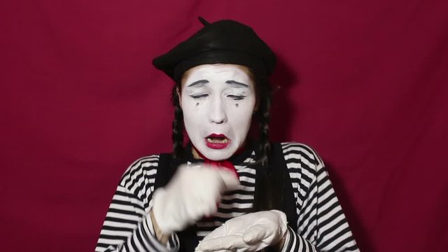 Beautiful mime girl depicts a strong upset and crying, looking at the camera. A beautiful mime girl in a striped shirt shows a sad face and tears. Portrait on a red background.