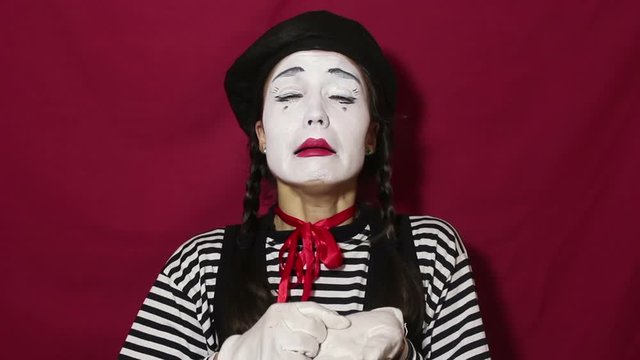 Beautiful mime girl depicts a strong upset and crying, looking at the camera. A beautiful mime girl in a striped shirt shows a sad face and tears. Portrait on a red background.