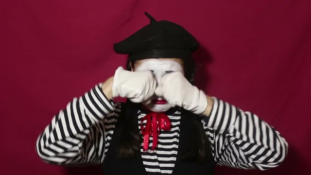Beautiful mime girl depicts a strong upset and crying, looking at the camera. A beautiful mime girl in a striped shirt shows a sad face and tears. Portrait on a red background.