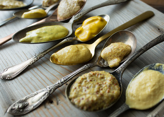 Several types of mustard shown in spoons on a wooden background. 