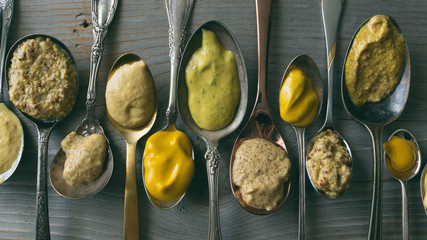 Several types of mustard shown in spoons on a wooden background. 