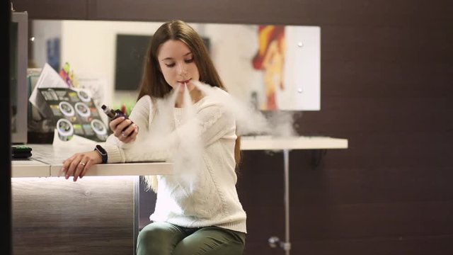 Vaping teenager. Young pretty white caucasian teenage girl with problem skin smoking an electronic cigarette indoors. Life-threatening bad habit. Vape activity.