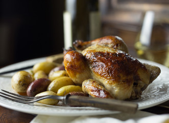 Side view of a roasted Cornish hen with potatoes and rustic fork, white wine in the background. 
