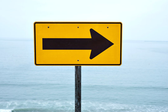 A yellow street sign with arrow pointing right at the beach with a view of the ocean