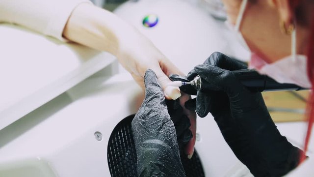 A manicurist in gloves polishes a client's nails with an apparatus before applying a gel coating