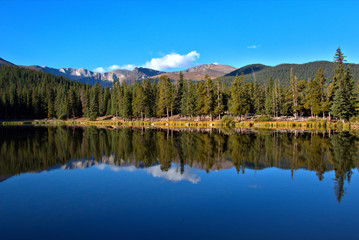 Breathtaking mountain lake with tall pine trees reflecting in the water with mountain peaks in the background with clear blue sky with puffy white clouds