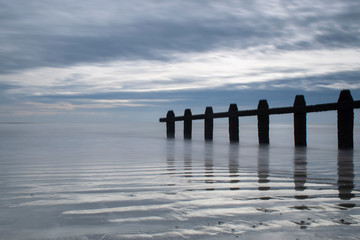 Reflections in the sand and the sea with dramatic cloud towards the sun, with a wooden groan reflecting in the water on the south coast of England.