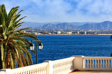 Beautiful seascape on the promenade, a street lamp under a palm tree looks at the sea, city and mountains in blur.