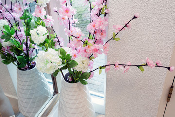 Bright pink and white colored fake flowers in a white vase near a window on a light day, plastic flowers in modern room