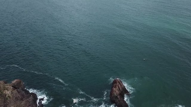 A flying camera takes a motorcyclist passing on a road near the sea with a view of the rocky shore.