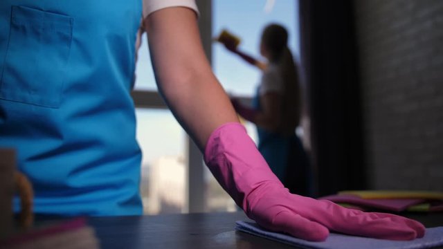 Closeup of female hand in glove thoroughly wiping table with duster after spraying detergent, blurred silhouette of workmate washing window. Skillful employees of cleaning service working in apartment
