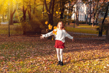 Beautiful little girl plays in the autumn park with yellow leaves on sunny day.