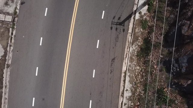 A flying camera takes a motorcyclist passing on a road near the sea with a view of the rocky shore.