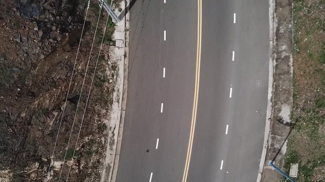 A flying camera takes a motorcyclist passing on a road near the sea with a view of the rocky shore.