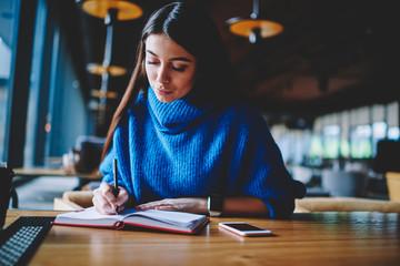 Attractive young talented female author notes new book articles while resting at cafeteria, thoughtful blogger girl casual dressed creating publication writing ideas in textbook at leisure indoors