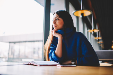 Beautiful young caucasian female model sitting and enjoying leisure indoors, attractive hipster girl 20 years old wearing in blue sweater dreaming while looking away and sitting at loft cafe
