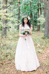 Obraz na płótnie Canvas The bride with wreath on head in a stylish wedding dress in nature forest with a rustic bouquet. Photo shoot in pine forest in the style of fine art.