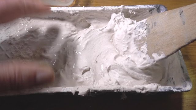 Close POV shot of a man’s hands adding some water to a small batch of freshly mixed plaster in a plastic tray, then slowly and carefully stirring it together with a wooden spatula.