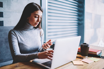 Skilled beautiful female graphic designer using smartphone for chatting with colleagues while sitting in favourite cafeteria and working on modern laptop computer, pensive woman checking email indoors