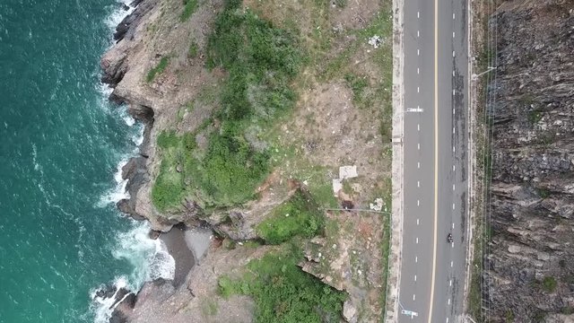 A flying camera takes a motorcyclist passing on a road near the sea with a view of the rocky shore.