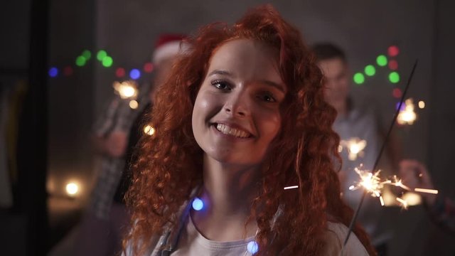 Cheerful curly redhaired girl with colorful lights on neck posing for camera - smiling, holding her bengal light while her friends dancing on the blurred background in decorated room. Friends