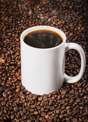 Steaming hot black coffee in plain white mug sitting on roasted coffee beans background