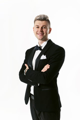Portrait of young smiling handsome man in tuxedo stylish black suit, studio shot isolated on white background. Showman or toastmaster in jacket with bowtie standing with cross hands