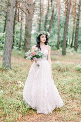 Fototapeta na wymiar Full-length view of the charming bride in greenery wreath with the wedding bouquet looking aside in the spring forest.