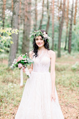 Obraz na płótnie Canvas beautiful bride walking in a coniferous forest in a wreath on her head and a luxurious wedding dress, holding rustic wedding bouquet