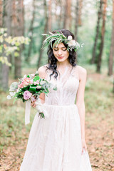 Obraz na płótnie Canvas Wedding ceremony in rustic style. Beautiful bride with green wreath on the head, with bouquet in her hand, posing in pine forest