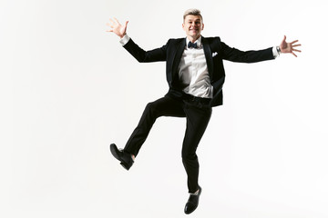Fototapeta na wymiar Portrait of young smiling handsome showman in tuxedo stylish black suit, studio shot jumping at white background. Businessman in jacket with bowtie