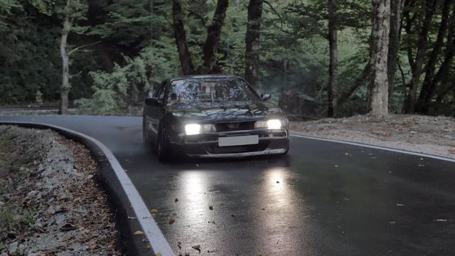 Black car starting drift with skidding and smoke on countryside road. Japanese car driving with slip and smoke on country highway on green forest trees background