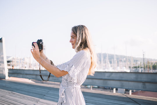 Side view of attractive woman in stylish sundress taking pictures via modern camera for photographing on leisure, charming hipster girl using equipment for making photos while travelling to Spain