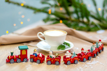 Delicious fresh festive morning cappuccino coffee in a ceramic white cup on the wooden table with decorative christmas train, red ornamentals, fireflies and spruce branches