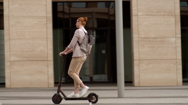 Following shot of young Caucasian woman with red hair wearing eyeglasses riding electric kick scooter in city center