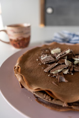 Сhocolate pancakes with chocolate and waffles on a pink plate and a cup of coffee on a white table. Vertical