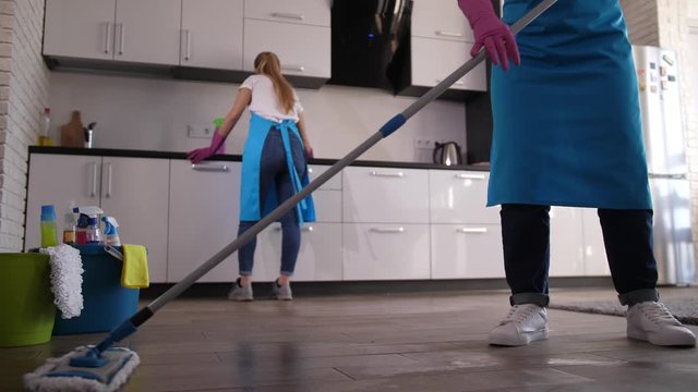 Professional cleaning lady using mop while washing floor, her workmate wiping kitchen surface on background. Two women, workers of cleaning service, in aprons and gloves during domestic clean up