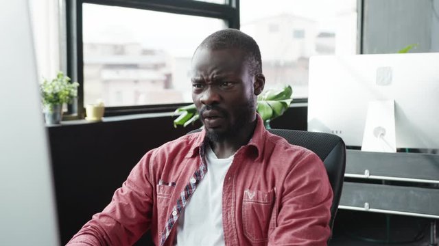 Black male employee sitting by desktop computer reading news screqing up face of disgust or disappointment working in company office.