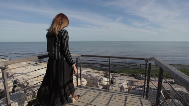 Woman with elegant dress moved by the wind watching the sea on the horizon leaning against a railing. Concept: wanderlust, travel lifestyle. Caorle, Venice, Italy
