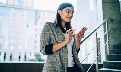 Smiling young woman sending message on smartphone standing on urban setting, positive trendy...
