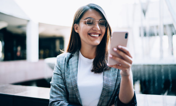Happy woman in eyewear smiling while watching trailer of future comedy connected to 4g wireless for browsing movie website, cheerful young blogger reading received message in group chat with followers
