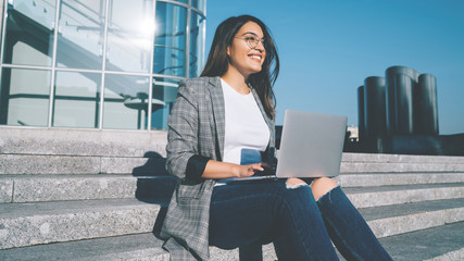 Happy remote copywriter looking away and smiling while sitting on urban setting with modern...