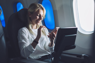 Smiling caucasian female passenger texting message on mobile phone while enjoying comfortable flight with Wireless network access on board. Young tourist woman choose airline with internet connection